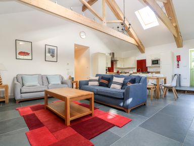 An open living area with two sofas a wooden coffee table a red rug exposed wooden ceiling beams skylight and a kitchen with dining table and chairs at Wheal Grey in Praa Sands
