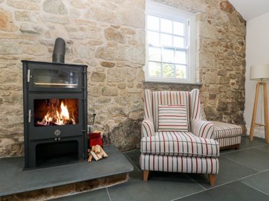A fireplace with a wood-burning stove next to a striped armchair and ottoman by a window in a room with a stone wall and tiled floor at Wheal Grey in Praa Sands