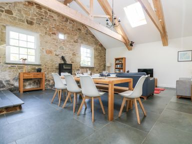 A dining area with a wooden table and white chairs next to a living room with sofas and a stone wall with windows at Wheal Grey in Praa Sands