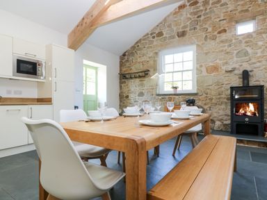 A dining area with a wooden table set with plates bowls and glasses surrounded by white chairs and a bench next to a stone wall with a window and a wood burning stove at Wheal Grey in Praa Sands