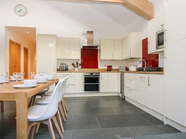 A kitchen with white cabinets a wooden dining table with white chairs and a red backsplash at Wheal Grey in Praa Sands