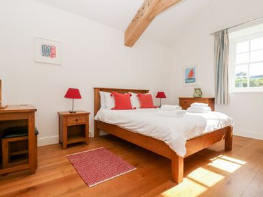 A bedroom with a wooden bed with red pillows and white linens two red lamps on wooden bedside tables a red patterned rug a wooden floor a wooden dresser and a window with striped curtains at Wheal Grey in Praa Sands