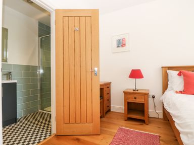 A bedroom with wooden furniture a red lamp a red cushion and an open door leading to a bathroom with green tiles at Wheal Grey in Praa Sands