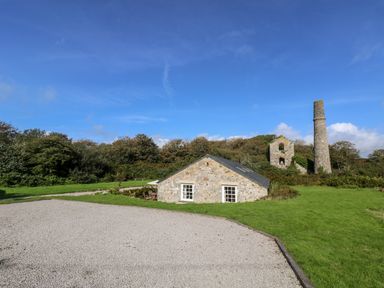 A stone building with white windows near a gravel area and tall chimney ruins surrounded by grass and trees at Wheal Grey in Praa Sands