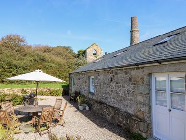 An outdoor patio with wooden chairs and table under a white umbrella beside a stone building with a chimney and a garden wall at Wheal Grey in Praa Sands