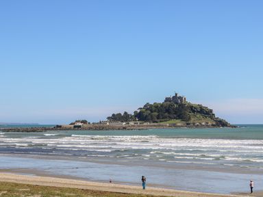 A beach with three people walking near the shore and a castle on a hill on an island in the sea at Wheal Grey in Praa Sands