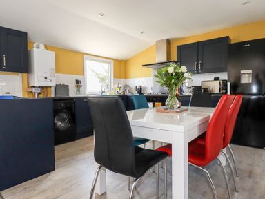 A kitchen with a white dining table surrounded by black red and blue chairs and a vase with flowers on the table at Kelyow in St Day