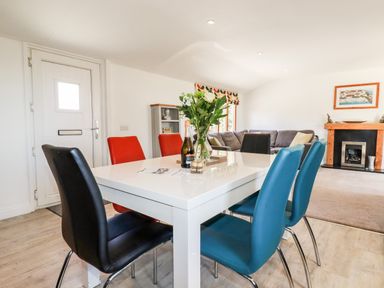A dining area with a white table surrounded by black red and blue chairs a vase with flowers bottles and glasses on the table a living area with a gray sofa and a fireplace in the background at Kelyow in St Day