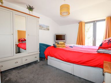 A bedroom with a bed covered by a red blanket a grey wardrobe a blue chair and orange curtains at Kelyow in St Day