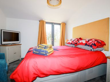 A bedroom with a red bedspread yellow headboard a television on a cabinet and curtains on a window at Kelyow in St Day