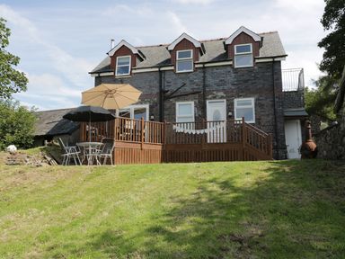 A two-story brick house with three dormer windows a wooden deck with two umbrellas and outdoor chairs on a grassy lawn at Hen Stabl in Llanrwst