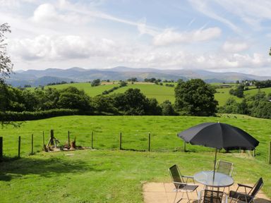 A grassy backyard with a glass table and chairs under a black umbrella overlooking green fields and distant hills at Hen Stabl in Llanrwst