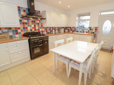 A kitchen with white cabinets a black stove a white dining table with four chairs and colorful tiled backsplash at Hen Stabl in Llanrwst