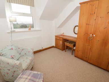A room with a floral armchair a wooden wardrobe a desk with a stool and a window at Hen Stabl in Llanrwst