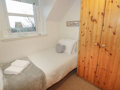 A small bedroom with a single bed white linens a grey heart shaped cushion wooden wardrobe and a window with a lamp at Hen Stabl in Llanrwst