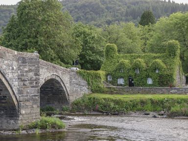 A stone bridge over a river with a house covered in ivy and trees in the background at Hen Stabl in Llanrwst