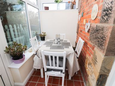 A dining area with a table covered by a tablecloth and four white chairs near windows and a brick wall at Corbett Cottage in New Haggerston near Holy Island