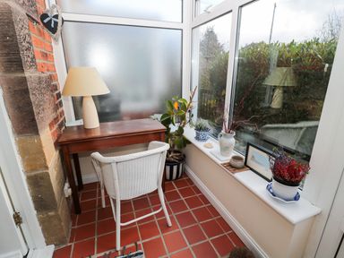 A small study area with a wooden desk, lamp, white chair, and potted plants next to frosted and clear windows at Corbett Cottage in New Haggerston near Holy Island