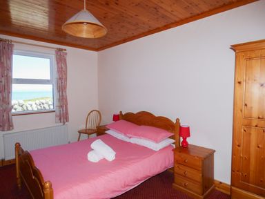 A bedroom with a wooden bed and wardrobe pink bedding red lamps and a window showing a stone wall and the sea at Seaview Cottage in Fanore County Clare