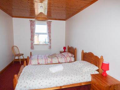 A bedroom with two wooden single beds bedside tables with red lamps a wooden chair a window with curtains and a wooden ceiling at Seaview Cottage in Fanore County Clare
