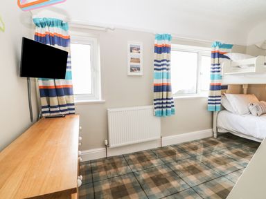 A small bedroom with bunk beds a wooden dresser wall-mounted television and two windows with striped curtains at The Beach House Criccieth