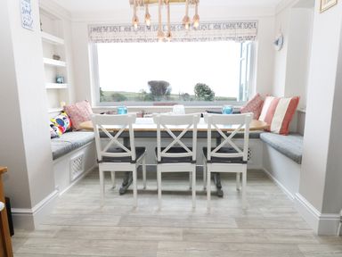 A dining area with a wooden table white chairs built in benches cushions and a window overlooking greenery at The Beach House Criccieth