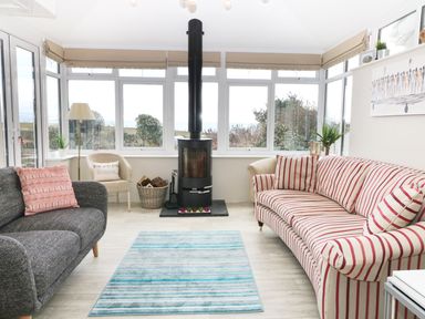 A sunroom with a black stove between a gray sofa and a striped sofa with a blue rug on the floor at The Beach House Criccieth