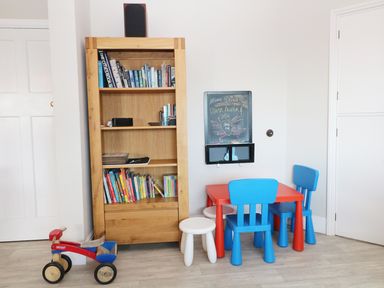 A wooden bookshelf with books a small red and blue table with two blue chairs and one white stool a small red and blue ride-on toy and a chalkboard on the wall at The Beach House Criccieth
