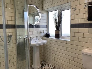 A bathroom with a pedestal sink glass shower door a window with a vase and rolled towel and patterned floor tiles at The Beach House Criccieth in Criccieth