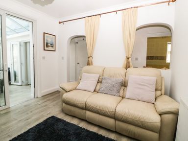 A room with a beige sofa with three cushions two beige and one gray a black rug on the floor light wood flooring and a glass door leading to another room at Pier View in Paighton