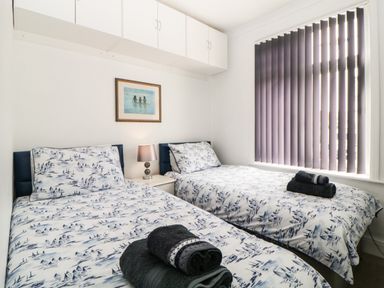 A bedroom with two single beds patterned bedding a nightstand with a lamp under wall cabinets and a window with vertical blinds at Pier View in Paighton