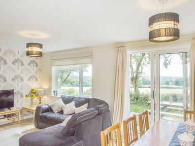 A living room with a black sofa table with sunflowers and a dining table near large windows and sliding doors at Dartmoor View in Gunnislake