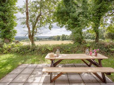 A wooden picnic table on a stone patio with trees and fields in the background at Dartmoor View in Gunnislake