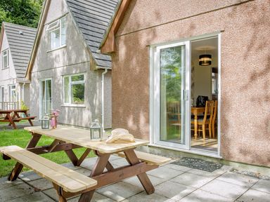 An outdoor patio area with a wooden picnic table and chairs next to a building with sliding glass doors at Dartmoor View in Gunnislake
