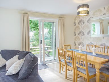 A dining area with wooden table and chairs next to a sliding glass door and a gray sofa with cushions at Dartmoor View in Gunnislake