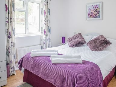 A bedroom with a double bed featuring purple cushions and a purple bedspread with white towels near a window with patterned curtains at Dartmoor View in Gunnislake