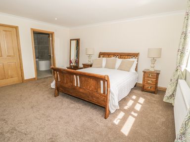 A bedroom with a wooden bed white bedding two bedside tables with lamps a standing mirror and an open door to a bathroom at Rede River Cottage West Woodburn