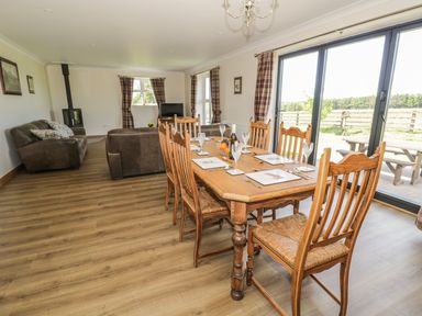 A dining area with a wooden table and chairs set for six next to sliding glass doors leading outside and a living area with sofas and a stove in Rede River Cottage in West Woodburn