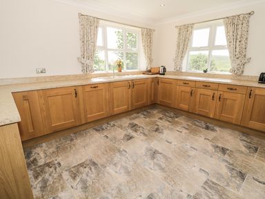 A kitchen with wooden cabinets granite countertops two windows with floral curtains a kettle and toaster on counters and stone tile flooring at Rede River Cottage in West Woodburn