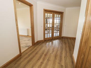A hallway with wooden flooring and door frames leading to a room with glass double doors and another room with a radiator at Rede River Cottage in West Woodburn