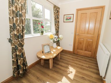 A small room with a wooden table holding flowers and a framed picture near a window with patterned curtains and a wooden door at Rede River Cottage in West Woodburn