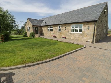 A stone house with a slate roof and green door next to a paved driveway and lawn at Rede River Cottage in West Woodburn