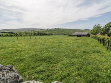 A fenced green field with scattered yellow flowers and a building in the distance at Rede River Cottage in West Woodburn