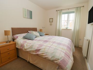 A bedroom with a double bed covered with a plaid duvet and cushions a wooden bedside table with a lamp a window with curtains and a wall-mounted television at 104 Sunrise Drive Filey