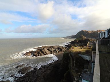 A rocky coastline with waves hitting the shore and buildings on the right at 10 Oceanside in Ilfracombe