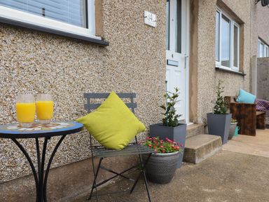 A small patio with a table holding two glasses of orange juice a chair with a green cushion plants in pots and a white door at Gwynfa in Rhyd Uchaf near Bala Gwynedd