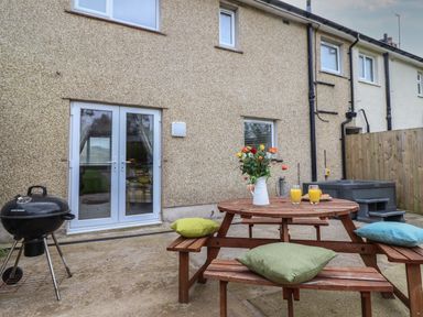 A backyard patio with a wooden picnic table with cushions and flowers hot tub next to a beige house with white doors and windows