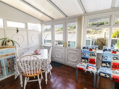 A dining area with a table covered by a floral tablecloth and four white chairs a cabinet a pair of patterned chairs and glass doors in a conservatory at Miners Cottage in Blaenau Ffestiniog
