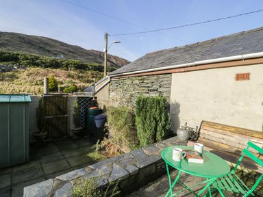 A small outdoor patio with a green table and chairs a wooden bench a book and mugs on the table at Miners Cottage in Blaenau Ffestiniog
