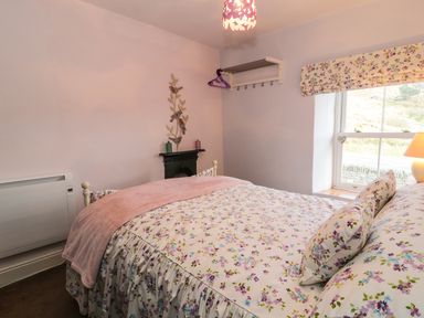 A bedroom with a floral bedspread a window with floral curtains a lamp and a small fireplace at Miners Cottage in Blaenau Ffestiniog